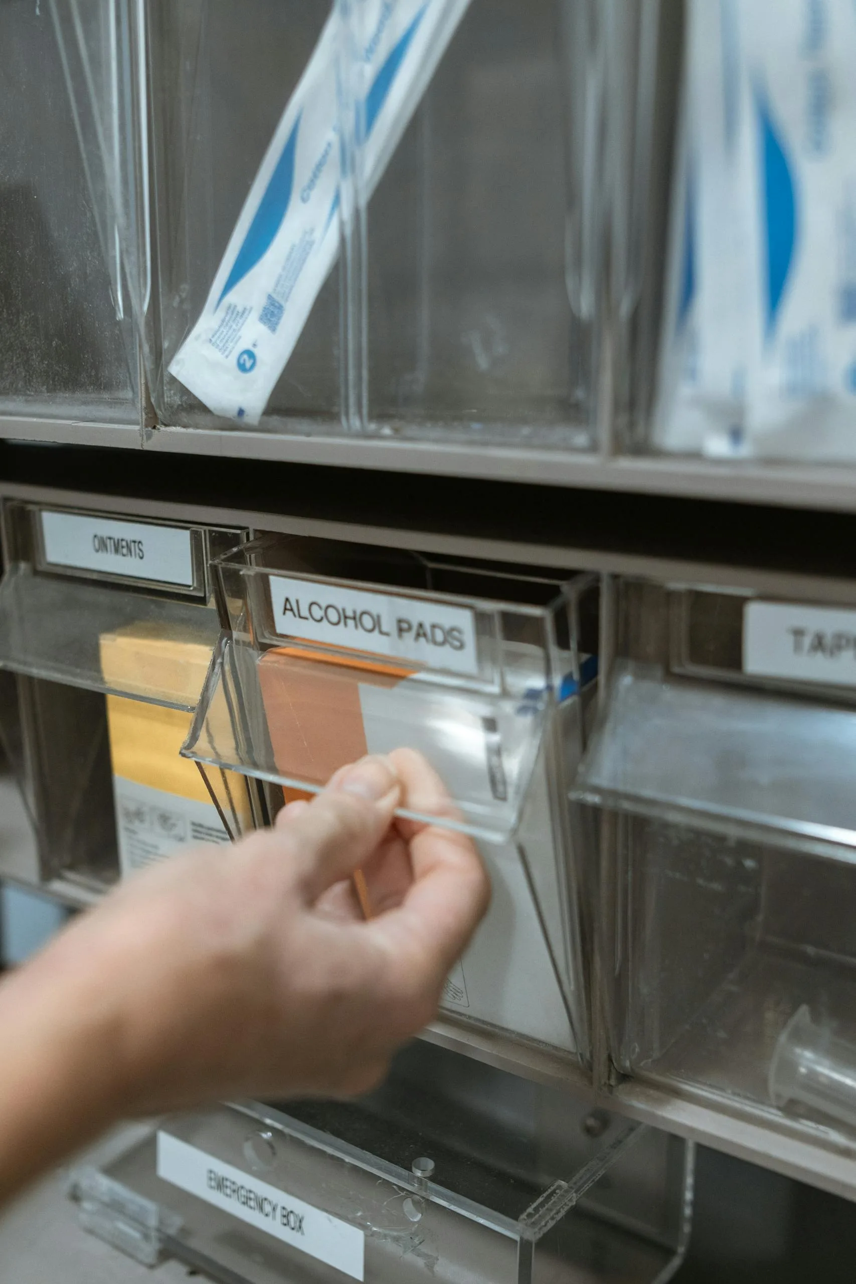 Detail of a hand retrieving alcohol pads from a medical supply dispenser on a shelf.